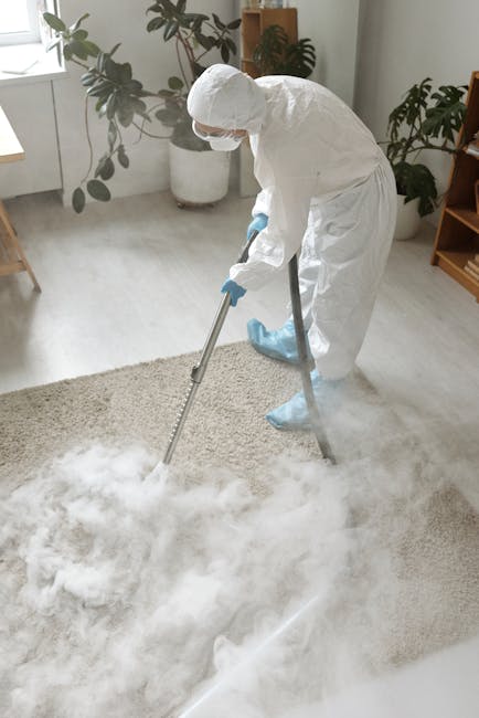 A person holding a yellow portable carpet cleaning or drying device with a black vent and control panel, positioned over a traditional patterned rug with floral motifs in shades of cream, beige, and m