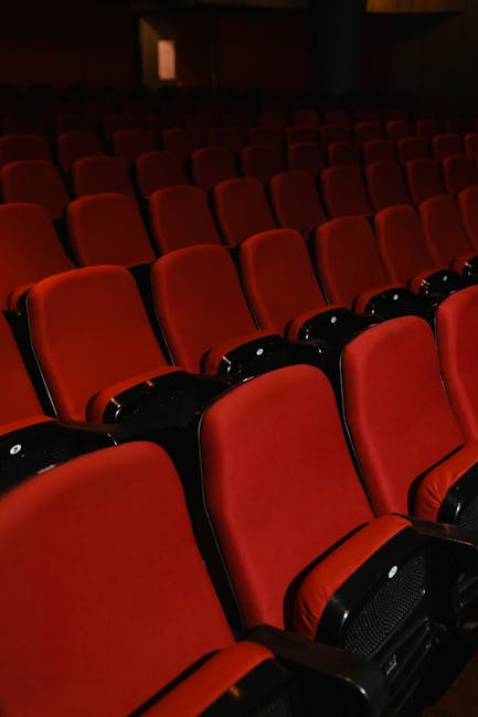 Close-up view of a row of black theatre chairs with red fabric seats and backrests, arranged on a dark wooden floor within a poorly lit space. The chairs are positioned on the left side of the image,