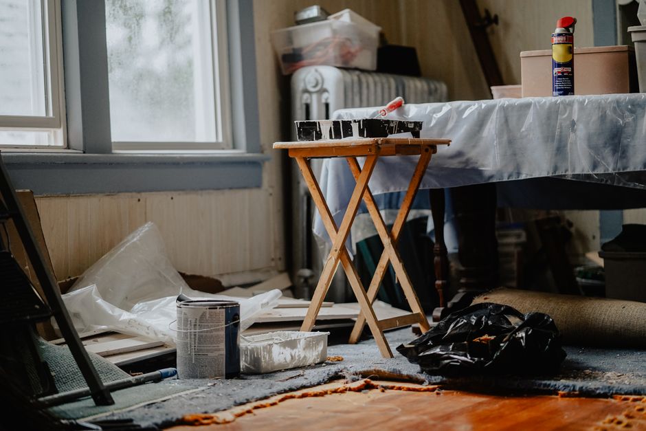An indoor space near a window with natural light illuminating a cluttered area, featuring a small wooden table topped with tools and materials for deep cleaning projects. The floor appears dusty with 